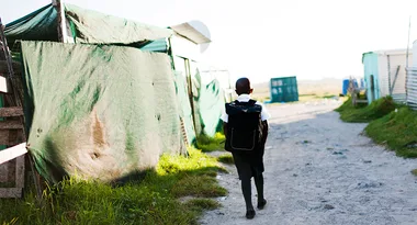 Boy walking alone on a dirt path with a school backpack, passing by makeshift structures covered with tarpaulin.