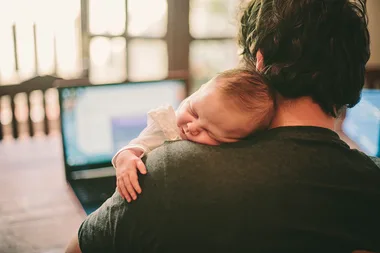 A person working on a laptop with a baby sleeping on their shoulder.