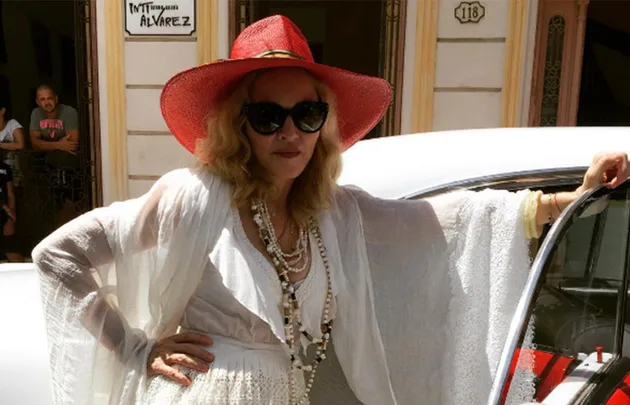 Woman in white dress, red hat, and sunglasses standing beside a car with a confident pose.