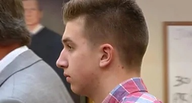Person in a courtroom wearing a red checkered shirt, with a blurred portrait in the background.