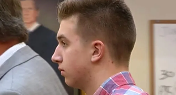 Person in a courtroom wearing a red checkered shirt, with a blurred portrait in the background.