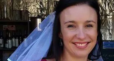 A woman smiling while wearing a bridal veil outdoors, with bottles on shelves in the background.