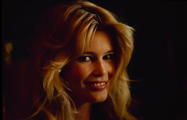 A woman with long blonde hair smiles softly in a dimly lit setting.