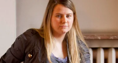 A person with long hair wearing a dark jacket and a slight smile, posed indoors against a beige background.