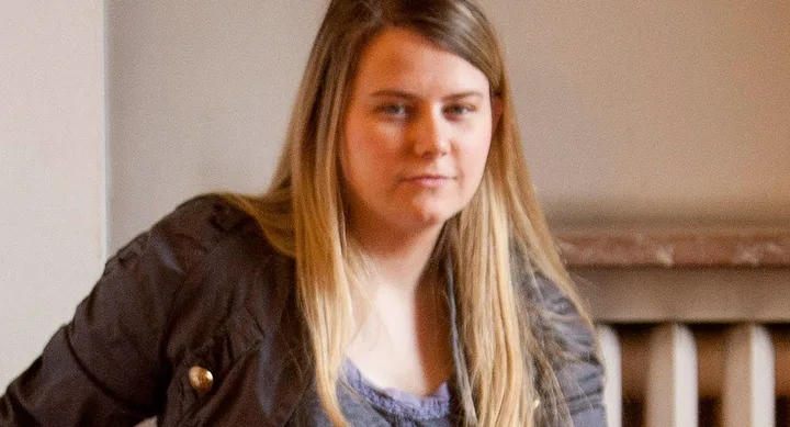 A person with long hair wearing a dark jacket and a slight smile, posed indoors against a beige background.