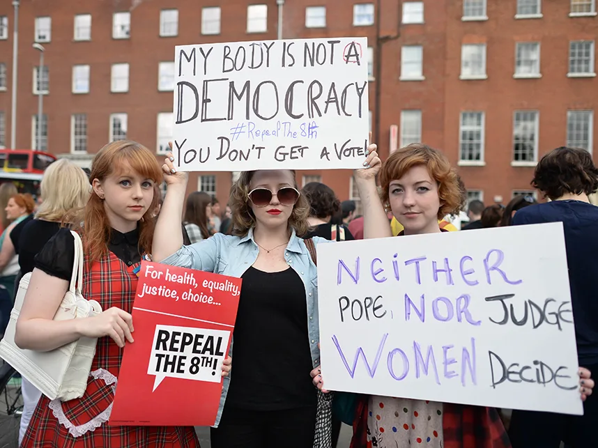 Three women at a protest hold signs supporting the repeal of the 8th amendment in a public square.