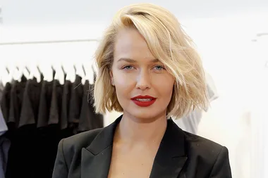 A person with short blonde hair and red lipstick wearing a black blazer, standing indoors.