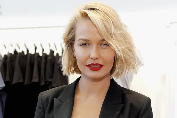 A person with short blonde hair and red lipstick wearing a black blazer, standing indoors.