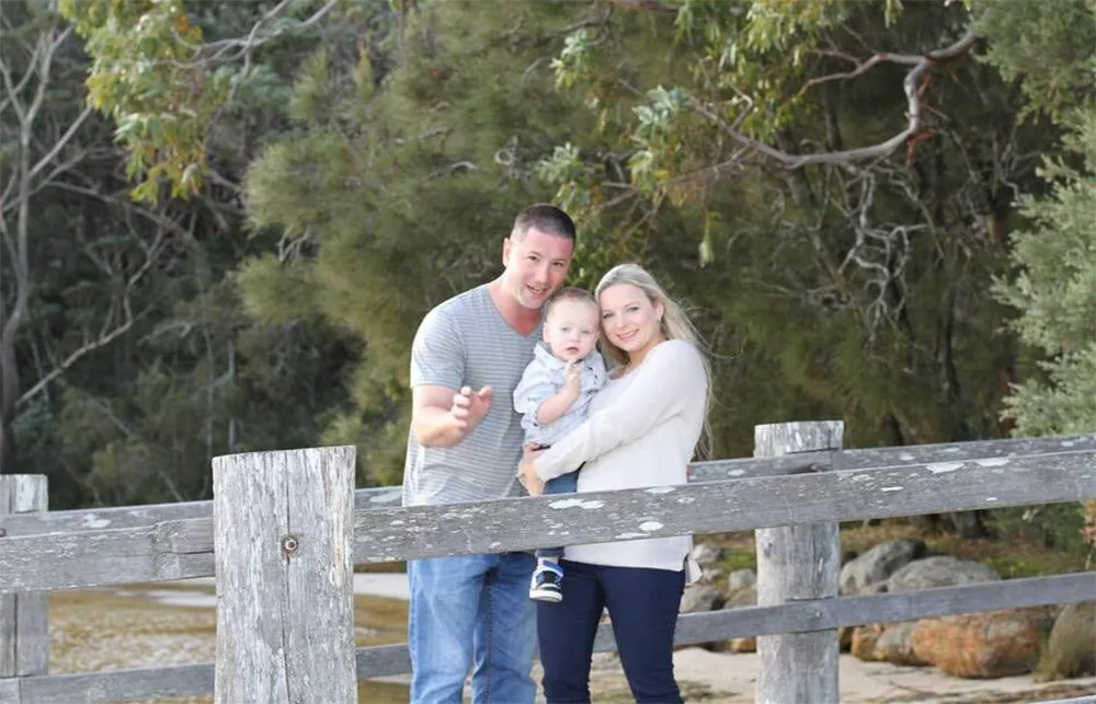 A family of three poses on a wooden bridge with trees in the background, the parents smile while holding their child.