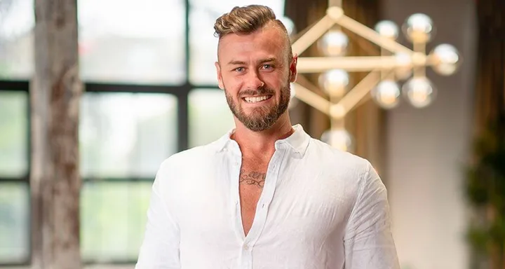 A man with a beard and styled hair smiles wearing a white shirt, standing indoors with a geometric light fixture in the background.