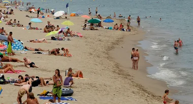 A busy beach with people sunbathing under umbrellas, swimming, and relaxing on the sand by the shoreline.