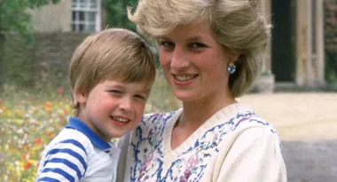 A smiling woman with short blonde hair holds a young boy, both looking at the camera in an outdoor setting.