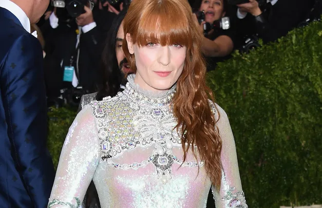 Red-haired woman in a shiny, embellished gown at a formal event, with photographers behind her.