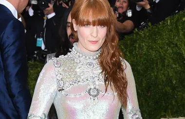 Red-haired woman in a shiny, embellished gown at a formal event, with photographers behind her.