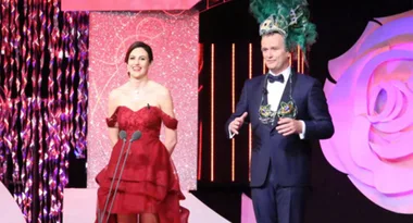 Two people on stage; one in a red gown, the other in a suit with a feathered headdress.