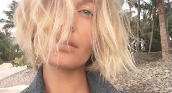 A person with blonde, tousled hair stands outside, partially obscured by hair, with palm trees in the background.