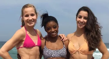 Three women in swimsuits smiling and posing on a beach with the ocean in the background.