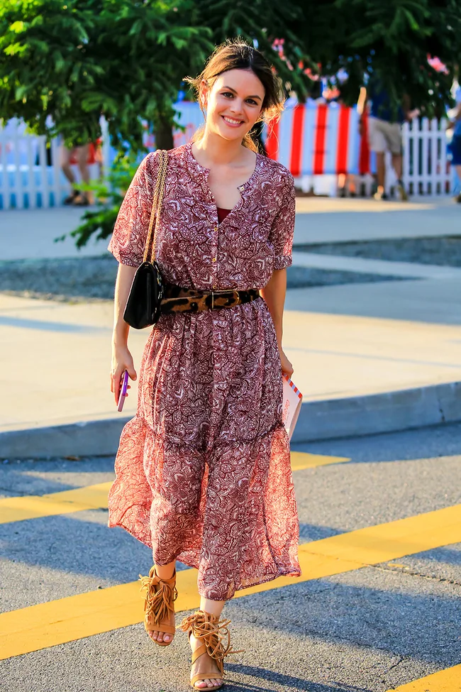 Young woman in a floral dress walking on a sunny street, holding a phone and a small striped item.
