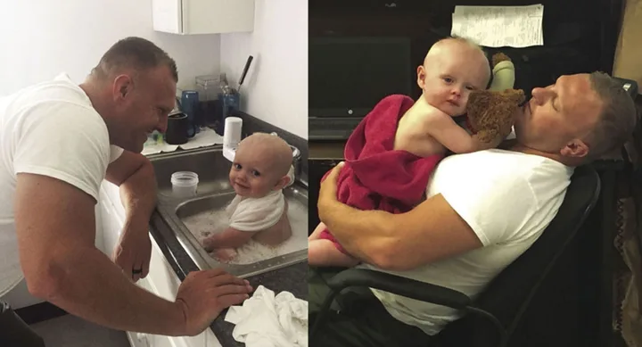 Man in white shirt interacts with baby in sink bath; cuddling baby wrapped in towel holding teddy bear on chair.