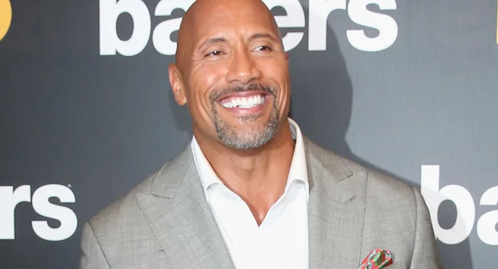 Man in a light gray suit smiling at a "Ballers" event backdrop.