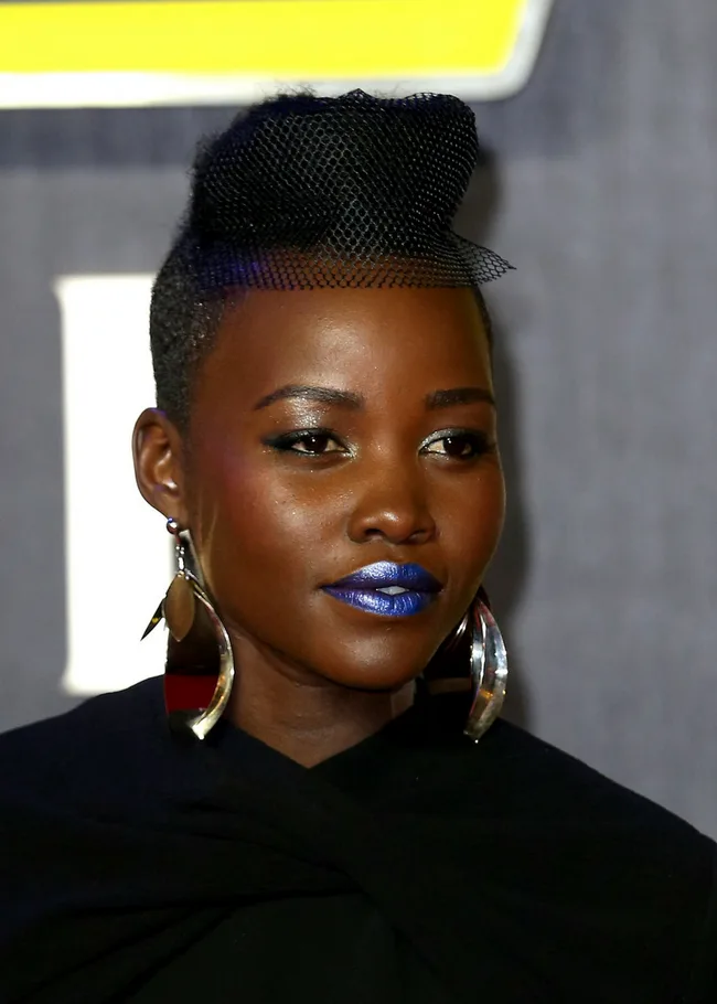 African woman with earrings and blue lipstick, wearing veil-like headpiece, attending an event.