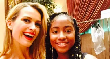 Two smiling women, one with blonde hair and red lips, and one with braided hair, in a warmly-lit indoor setting.