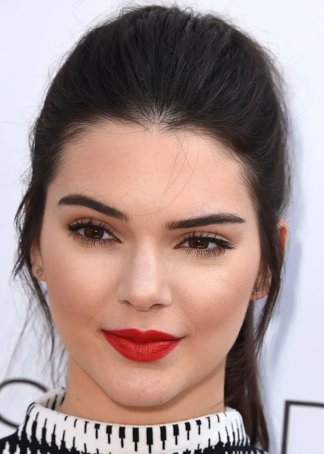Young woman with dark hair tied back, wearing red lipstick and a black and white patterned outfit, looking into the camera.