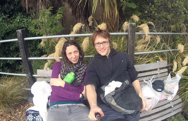 Couple sits on a park bench surrounded by bags, smiling at the camera on a sunny day.