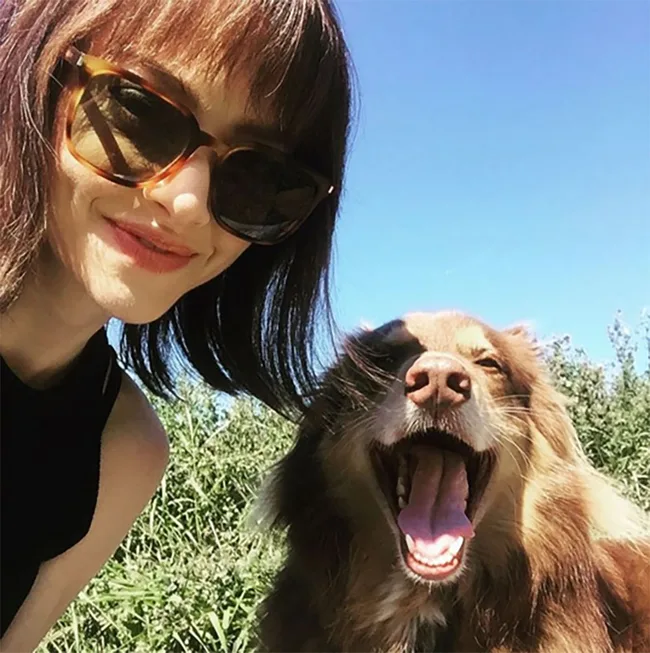 Woman in sunglasses and a yawning brown dog in a sunny outdoor setting.
