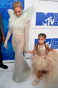 Woman in a sparkling, feathered gown holding hands with a young girl in a gold dress at an event.