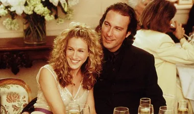 A smiling woman with curly blonde hair sits next to a man in a suit at a formal event, with flowers in the background.