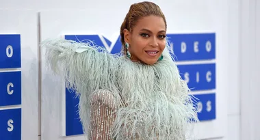 A person in an intricate feathered outfit poses at an event with blue and white signage in the background.