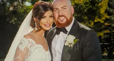 Bride in a white lace gown and veil, standing beside groom in a suit with boutonniere, outdoors.
