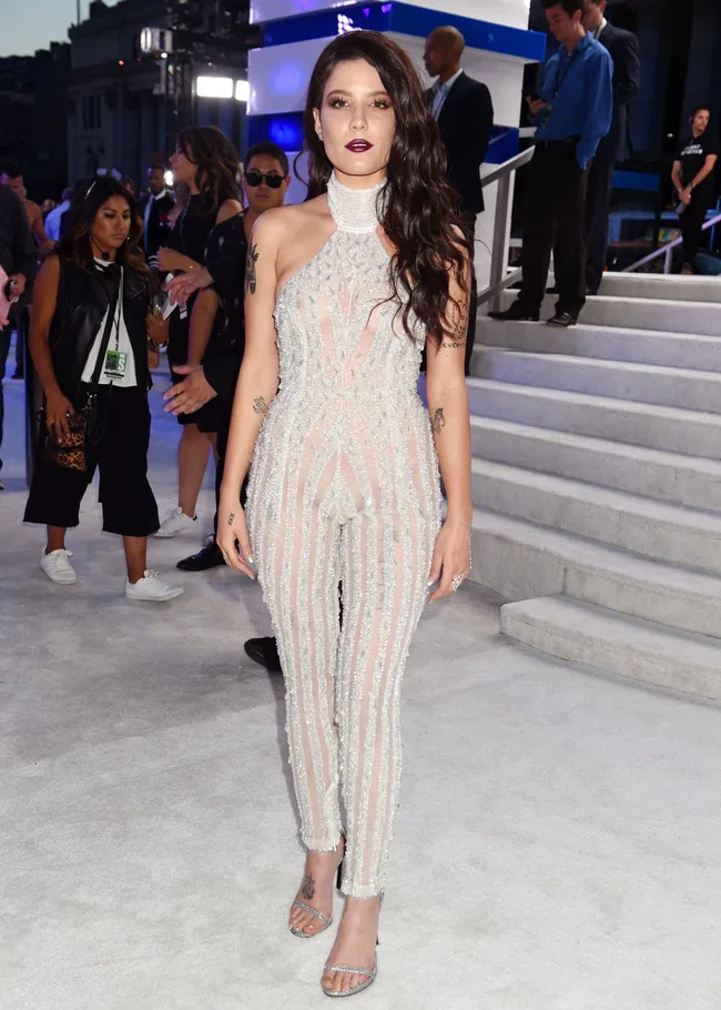 A woman in a sheer, sparkling outfit poses on a light carpet with people and stairs in the background.