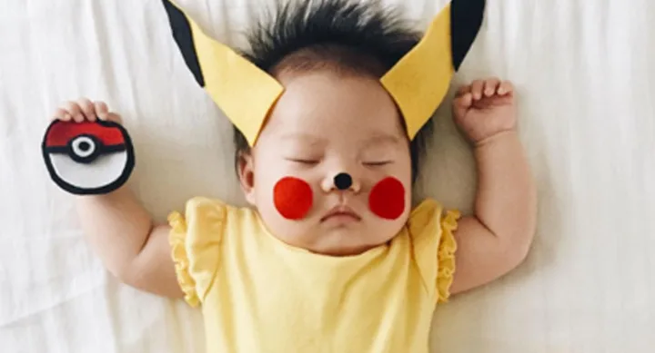 Baby in Pikachu costume asleep on a bed, holding a Poké Ball; wearing yellow outfit with ears and cheeks painted red.