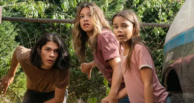 Three people, crouching and looking worried, are behind a fence with green foliage and beside an old vehicle.