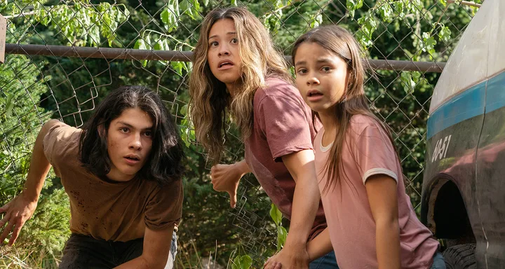 Three people, crouching and looking worried, are behind a fence with green foliage and beside an old vehicle.