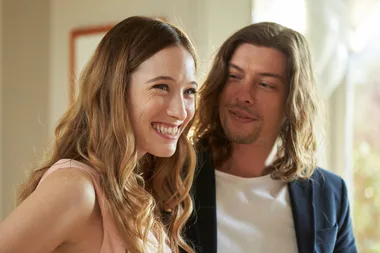 A happy woman with long brown hair smiles broadly, standing beside a man with long hair and a light beard indoors.