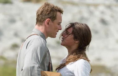 A man and a woman stand closely facing each other outdoors, with the man holding a wooden object.