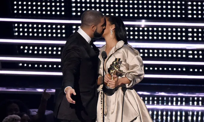 Two individuals embrace on stage with one holding a gold statue trophy; bright lights in the background.