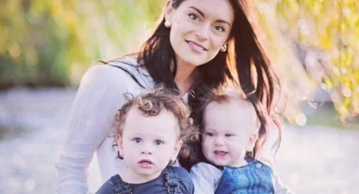 A woman with long dark hair smiles with two young children outdoors, surrounded by blurred greenery.