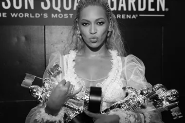 A person holding multiple awards stands in front of a Madison Square Garden sign, wearing a sheer, decorated outfit.