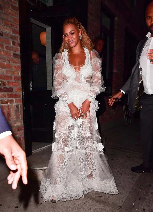 A woman in a sheer, white lace dress smiles while leaving a building at night.