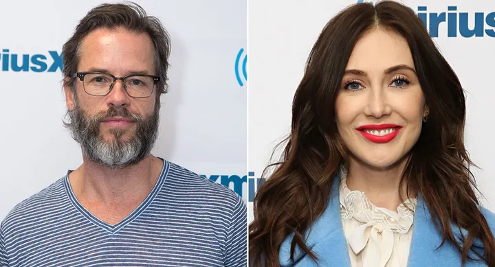 Male with glasses and beard in striped shirt; female with long hair in blue blazer, red lipstick smiling at event.