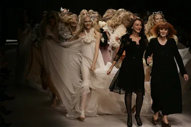 Models and designers walk down the runway in flowing dresses at a fashion show finale.