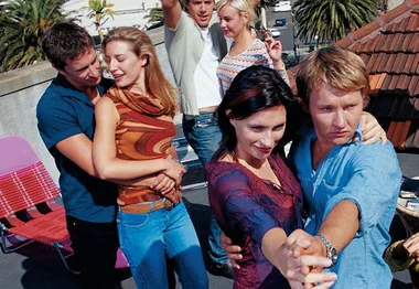 Six people dance on a rooftop, with two couples in the foreground and two people in the background near palm trees.