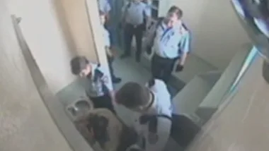 A CCTV image showing multiple officers in a small room with a person on the floor near a toilet, viewed from above.