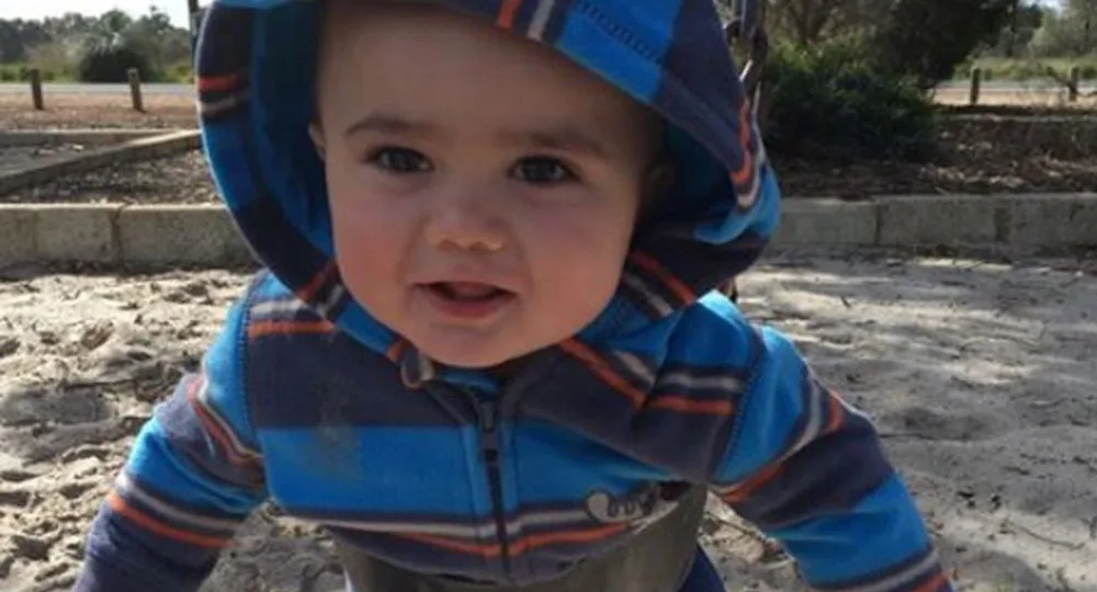 A toddler in a striped hoodie plays on a sandy playground, smiling at the camera.