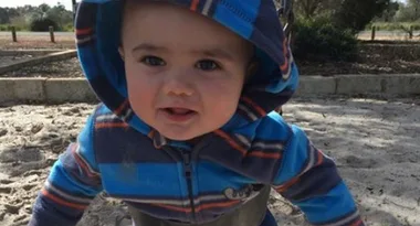 A toddler in a striped hoodie plays on a sandy playground, smiling at the camera.