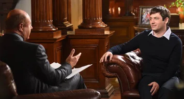 Two people sitting in leather chairs having a conversation in a warmly lit room with wooden decor and framed photos.